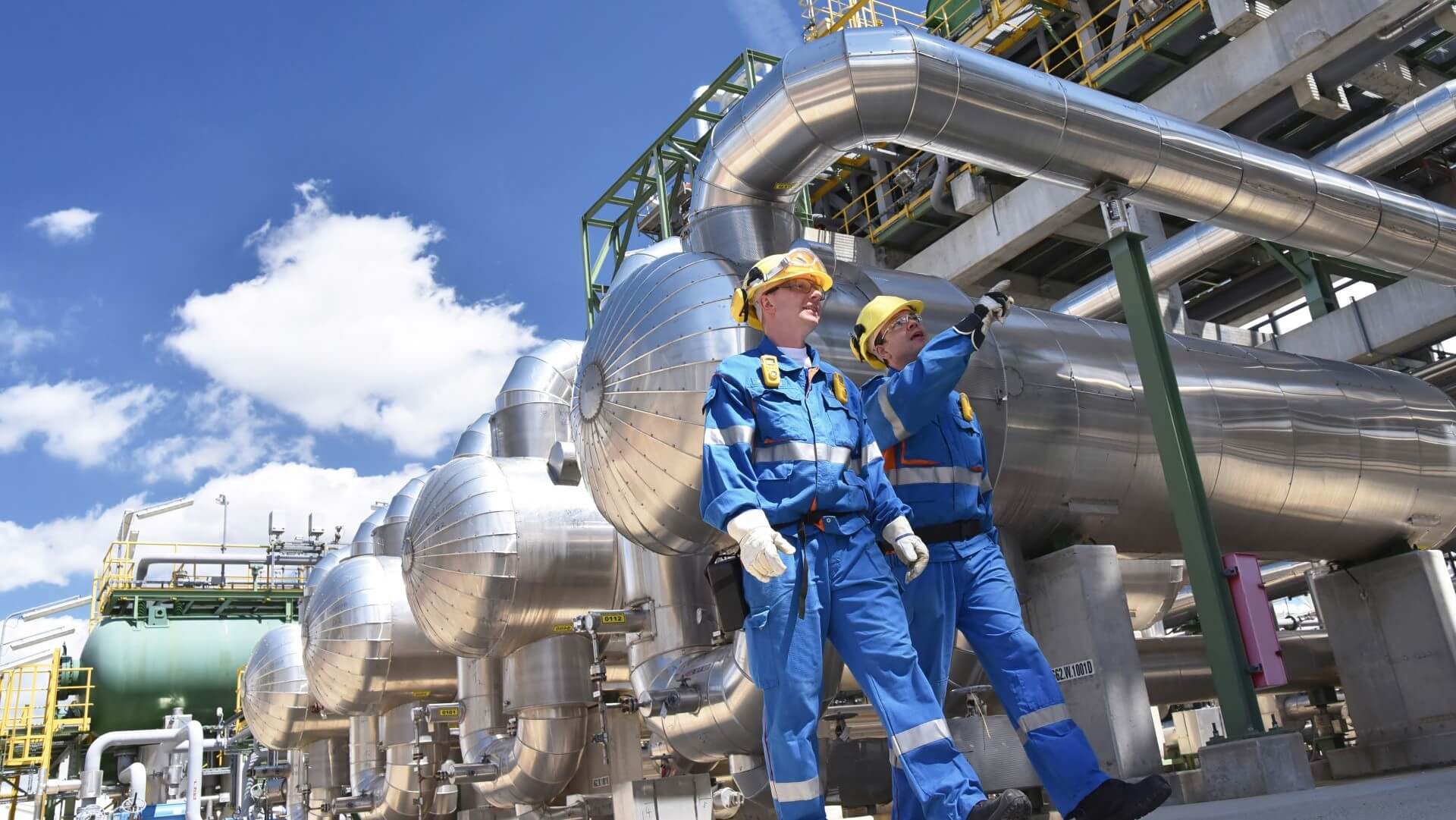 Two male engineers in boiler suits and hard hats walking and talking in front of pipework at a processing facility