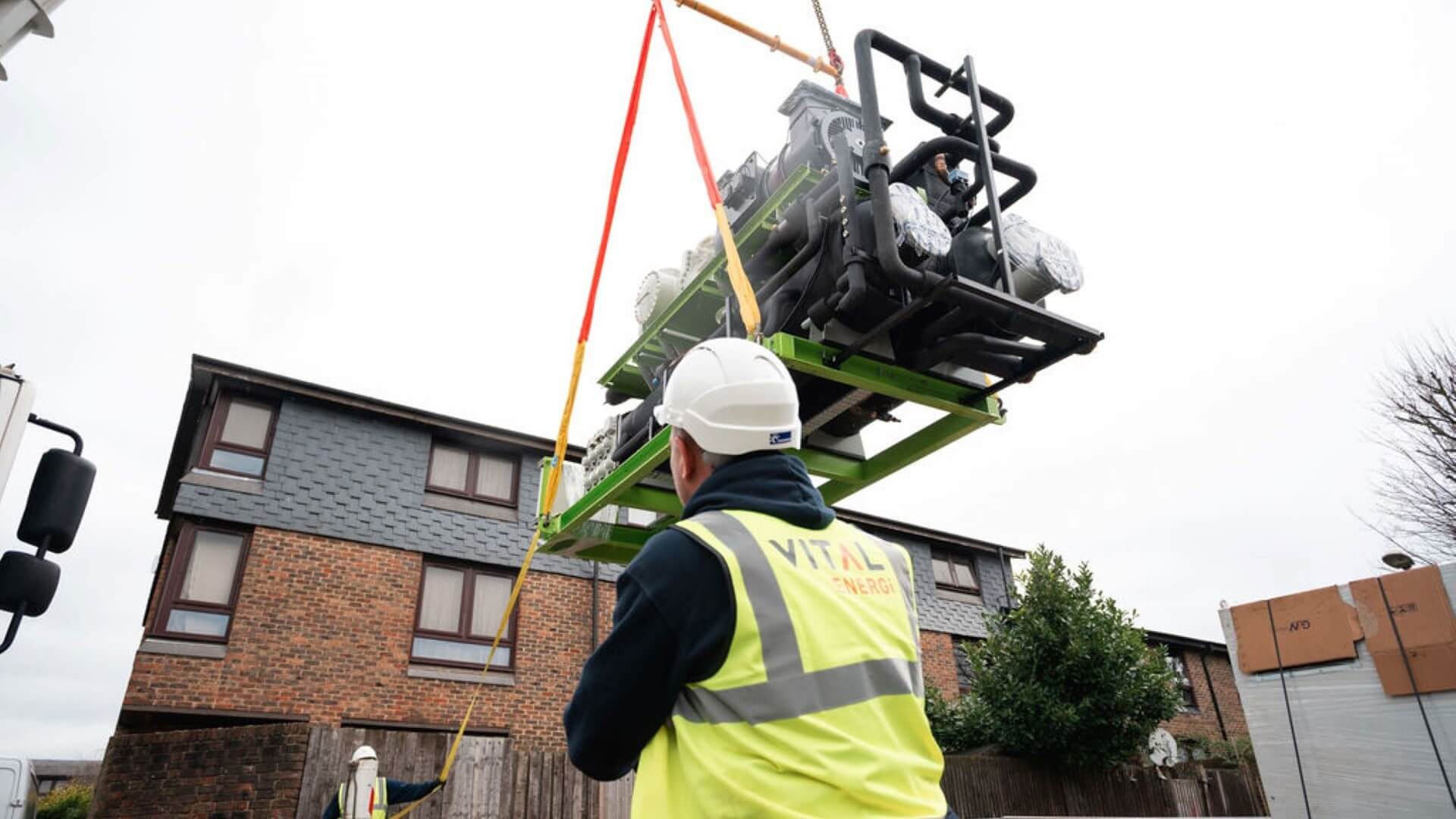 Man with hard hat and hi-viz jacket overseeing installation of heat pump being craned in, house in background