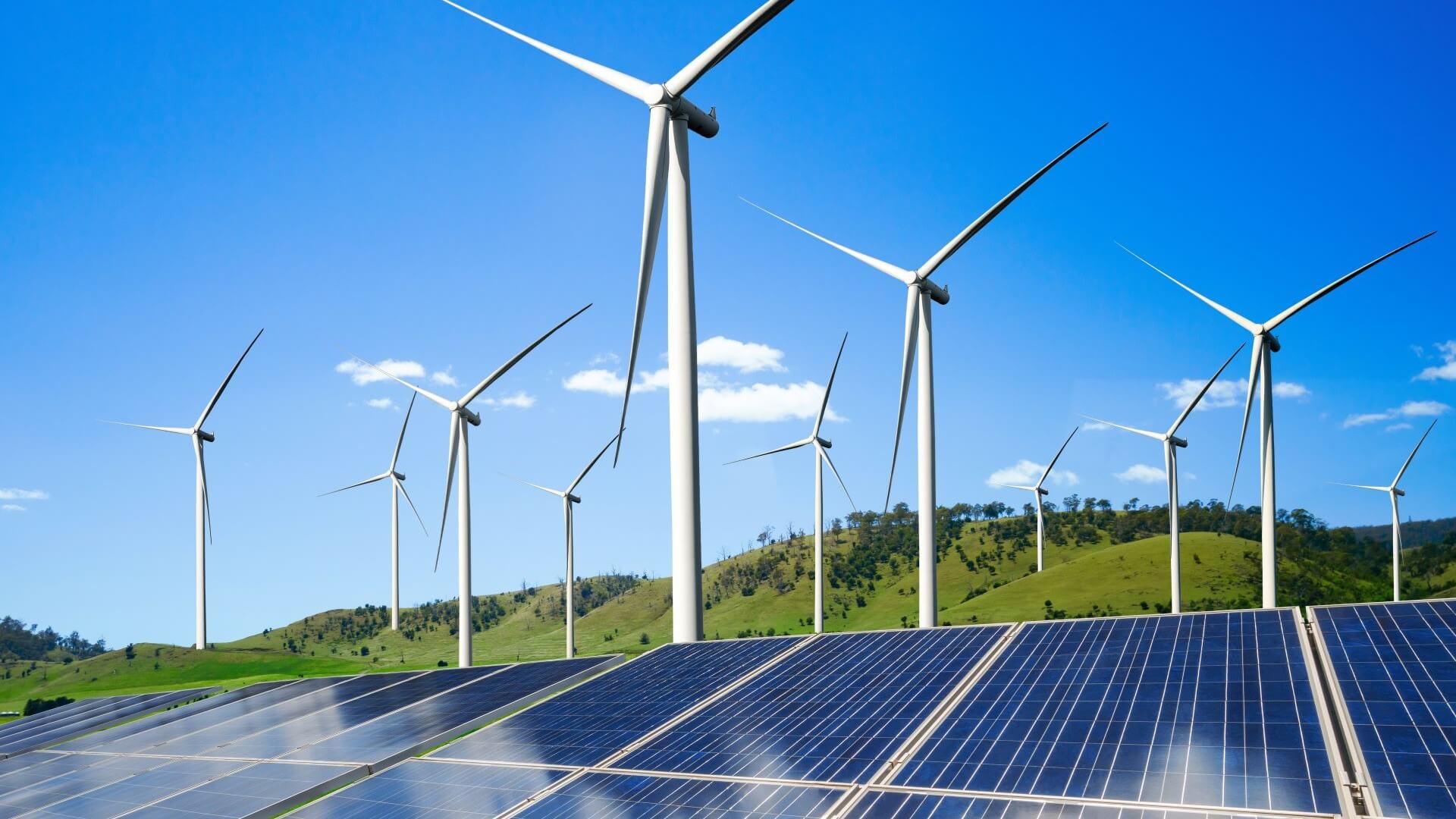 Row of solar panels in foreground and wind turbines in backgroud against green hills and blue sky