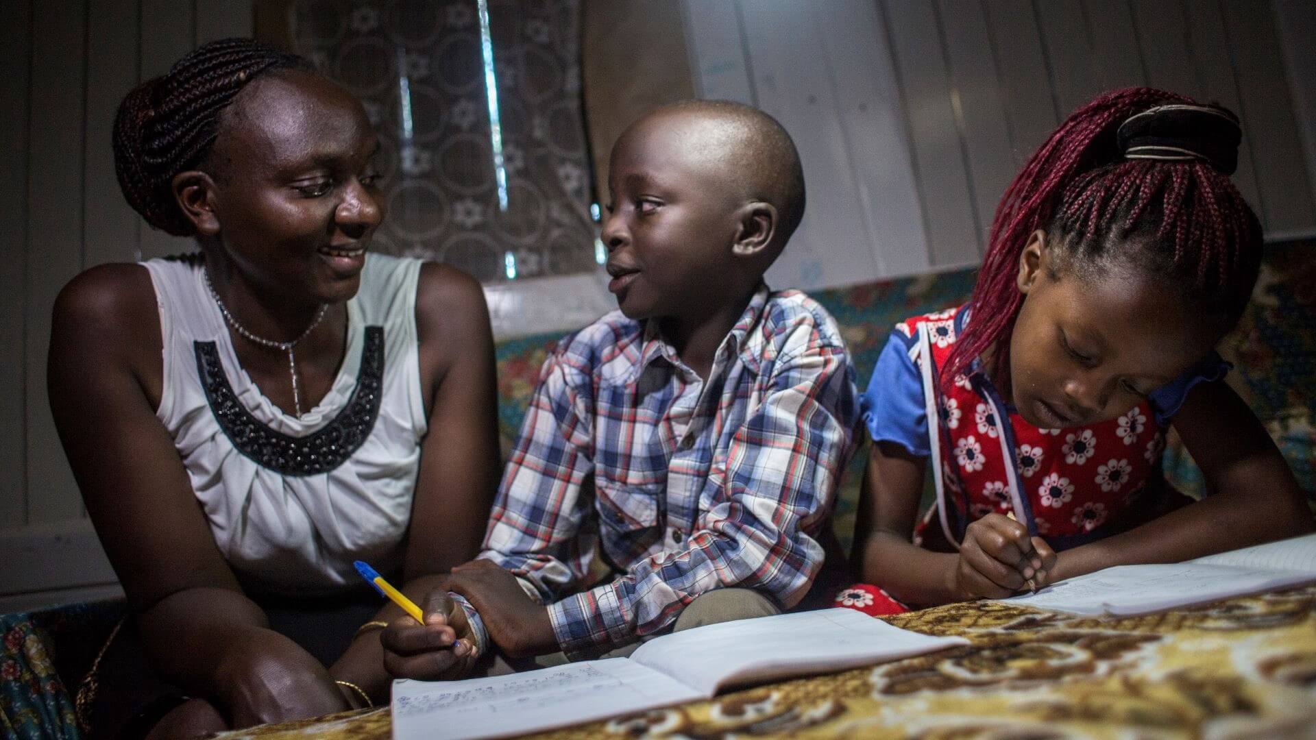 African woman sitting at table alongside two children writing out their homework in exercise books in dimly lit room