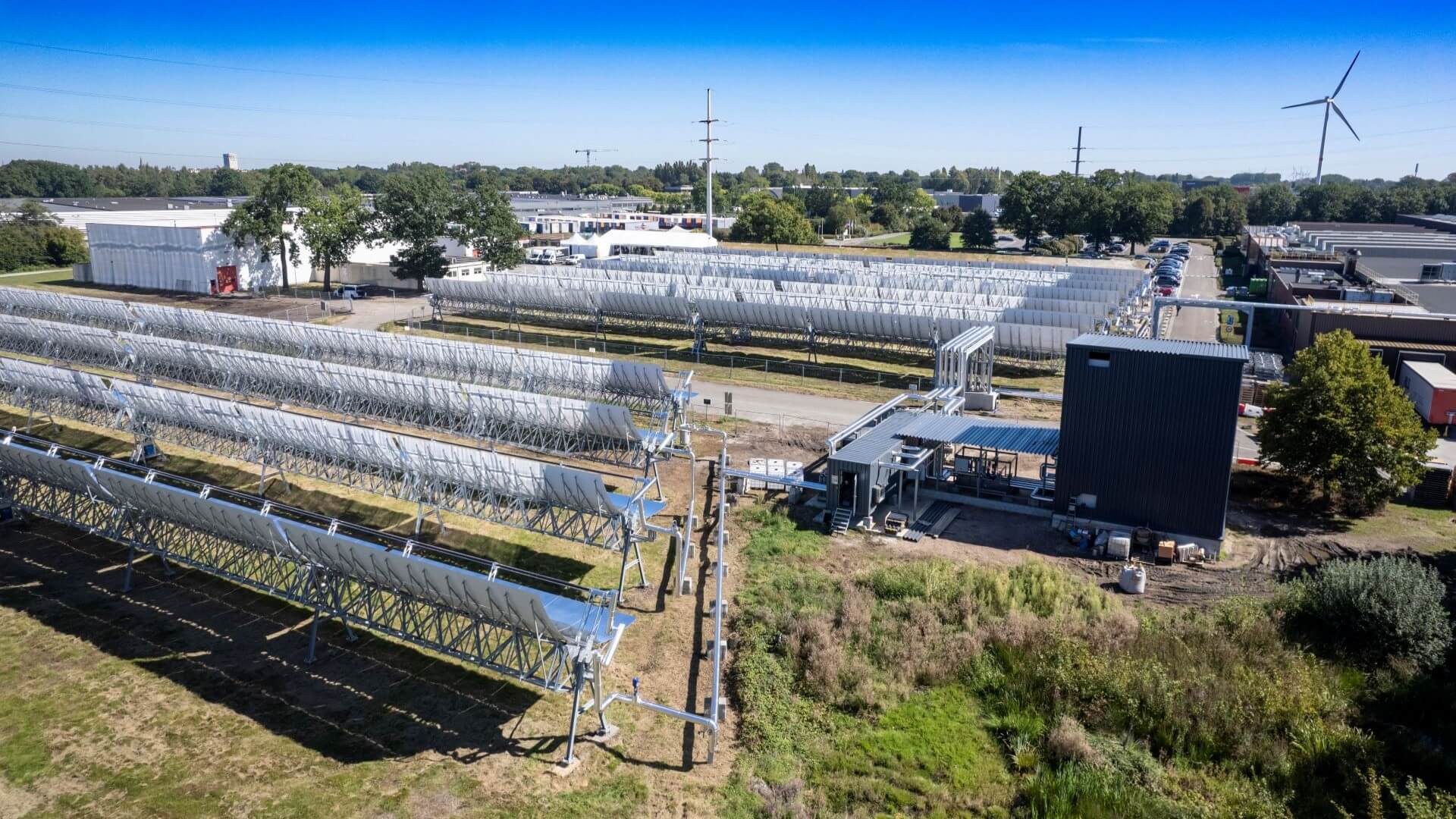 Overview of rows of parabolic mirrors at solar thermal installation with wind turbines on horizon in background