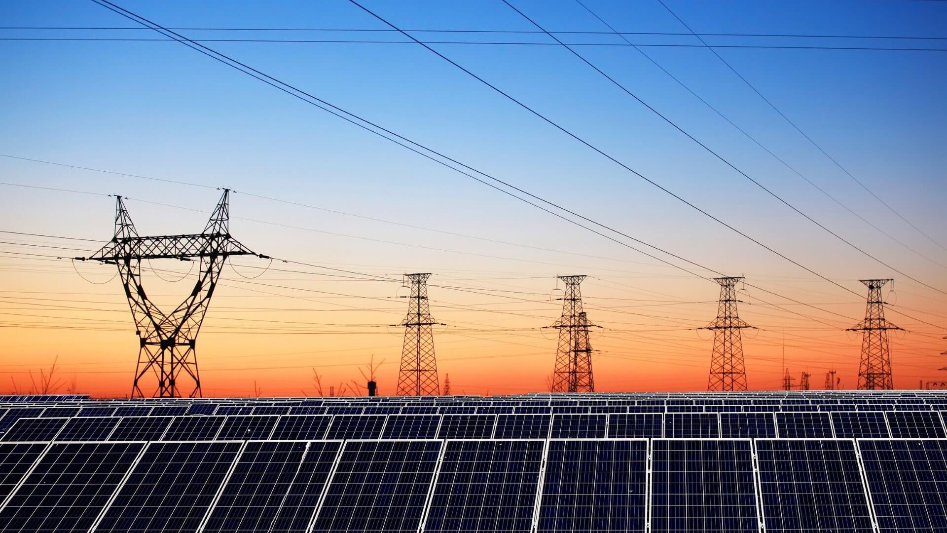 Line of solar panels in foreground with electricity pylons and transmission lines behind, set against a sunset