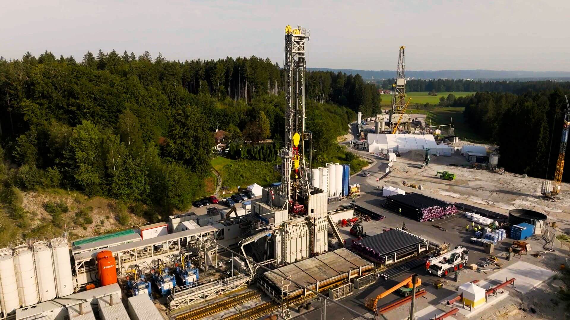 Aerial overview of geothermal project installation with industrial buildings and cranes in foreground and forest behind