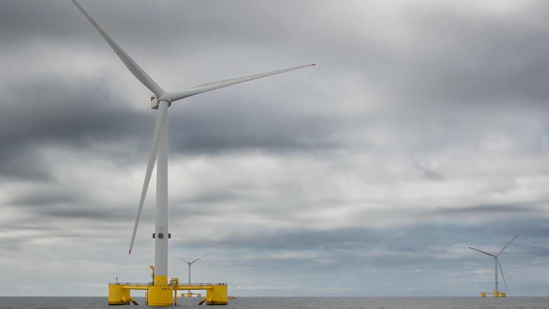 Floating offshore wind turbine in grey calm sea, in foreground set against grey sky, with another wind turbine in distant horizon