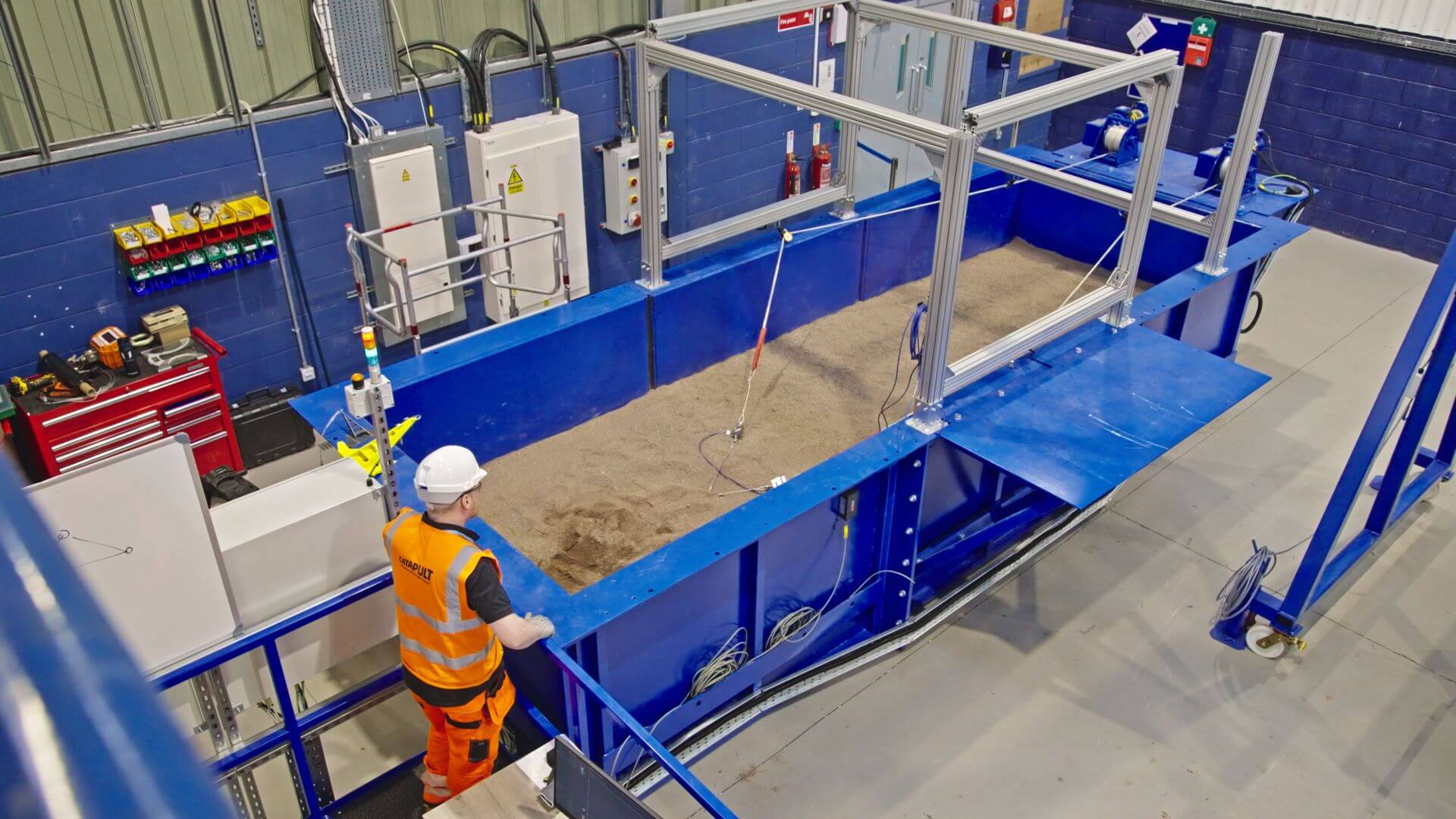 Man in orange high viz clothing and white hard hat standing next to large blue metal rectangular box filled with sand and surrounded by pipework and equipment