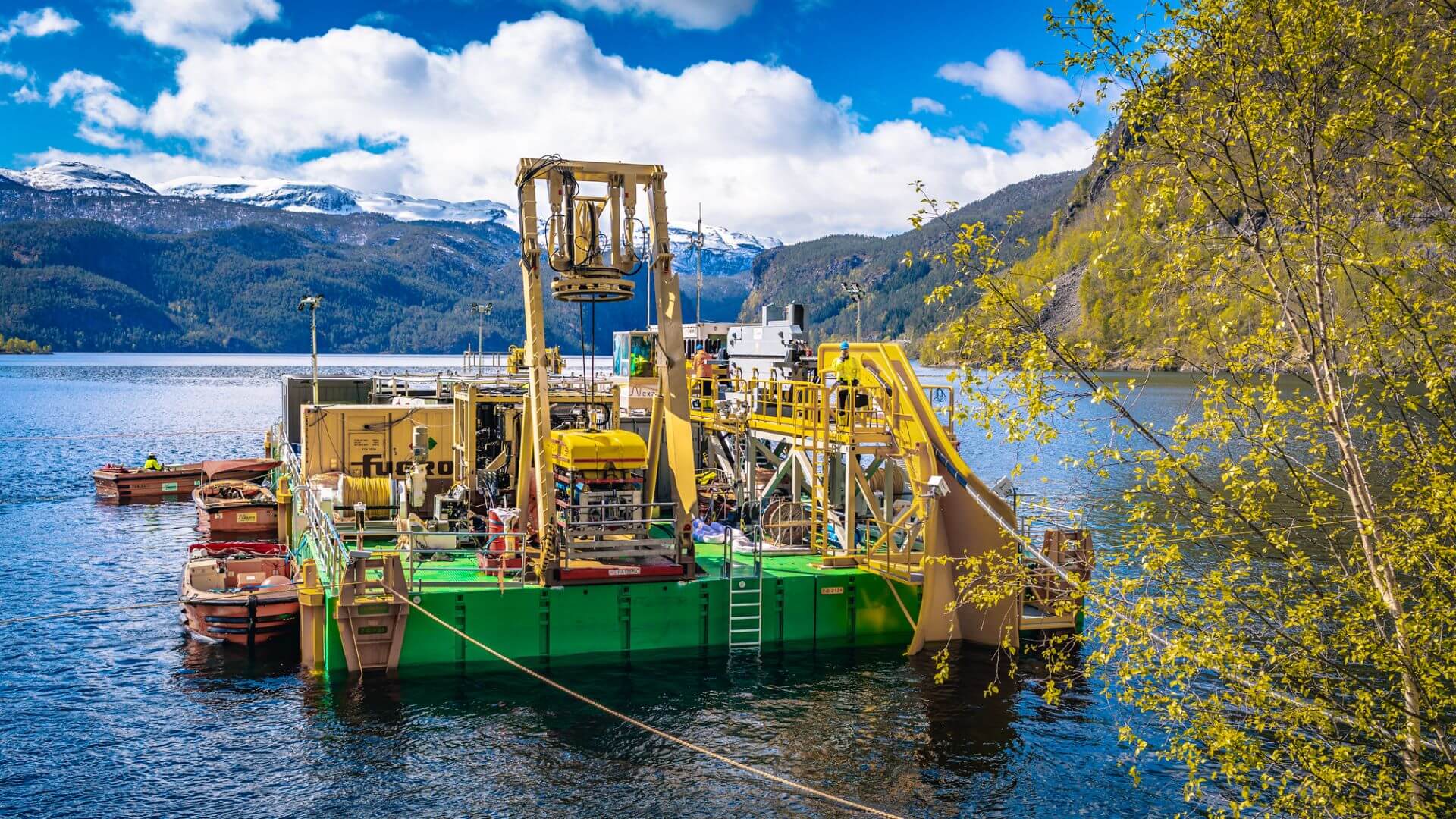 Barge with yellow equipment for installing pipes floating in calm water with hills in background