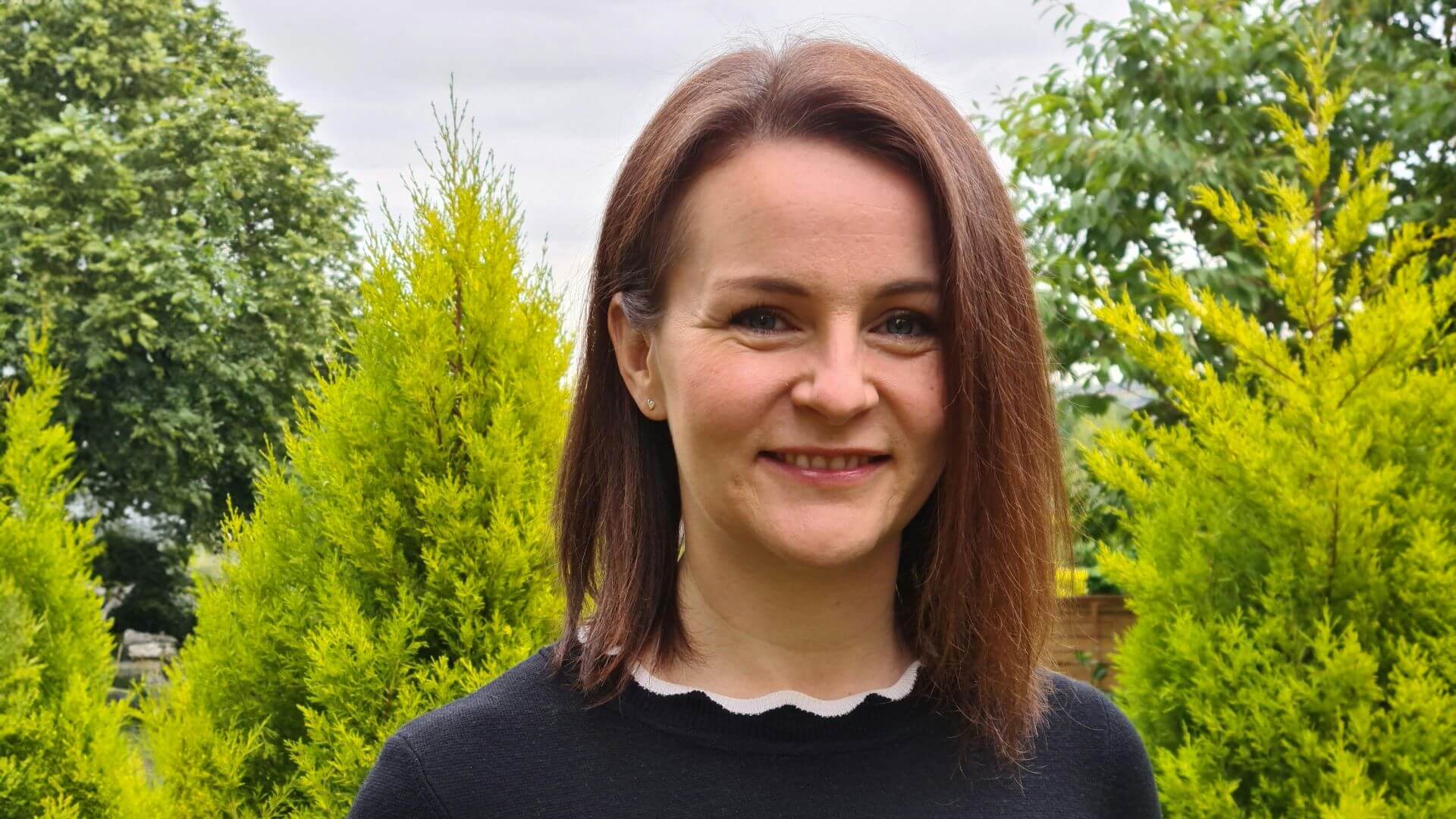 Head and shoulders photo of Emma Bridge standing in front of green conifers and leafy trees