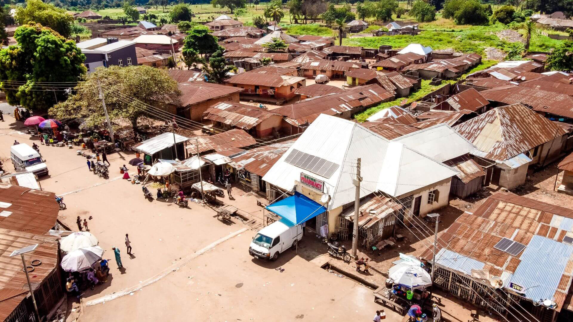 Group of rural African buildings, some with solar panels on roof, with a few vehicles and people milling about