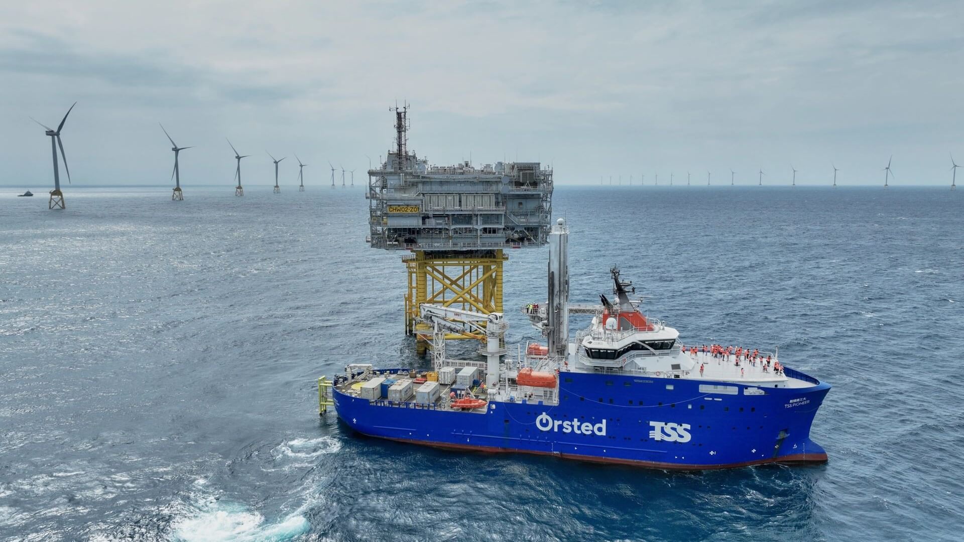 Vessel in foreground of photo, in calm sea, next to a small offshore platform structure and with rows of floating offshore wind turbines on horizon