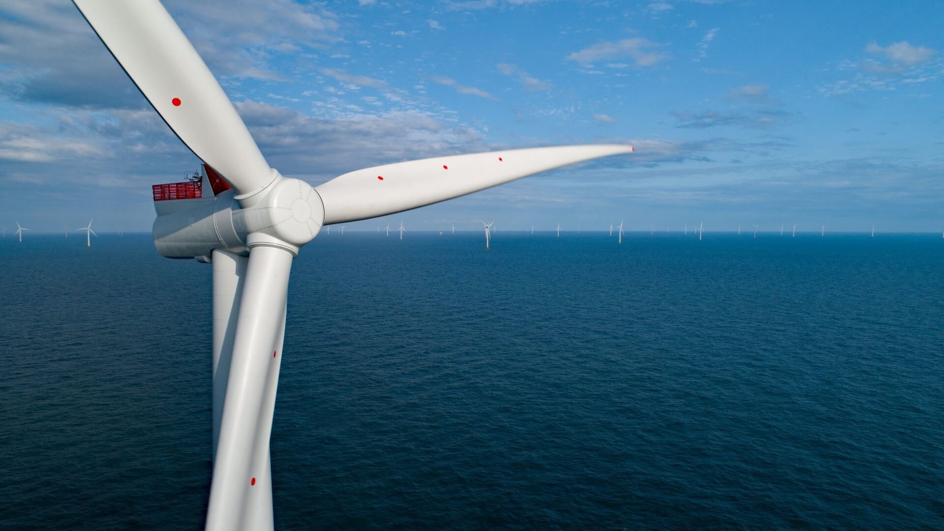 Close up view of nacelle and blades at top of offshore wind turbine in foreground, with blue sky and calm sea behind