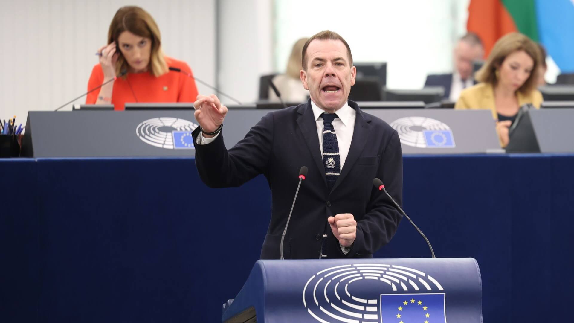 Man standing at lecturn, gesturing with hand and speaking, with men and women sat in background, listening to translations of speech