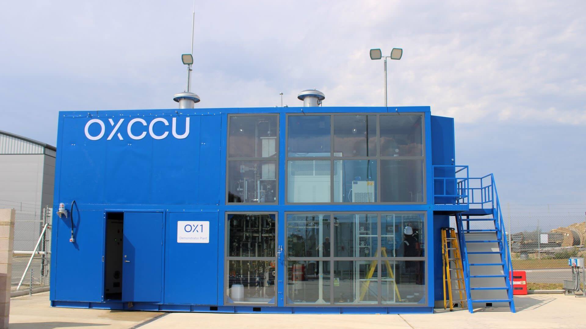 Small blue metal rectangular building, half the front of which is glass panelling, with metal staircase running up right hand side