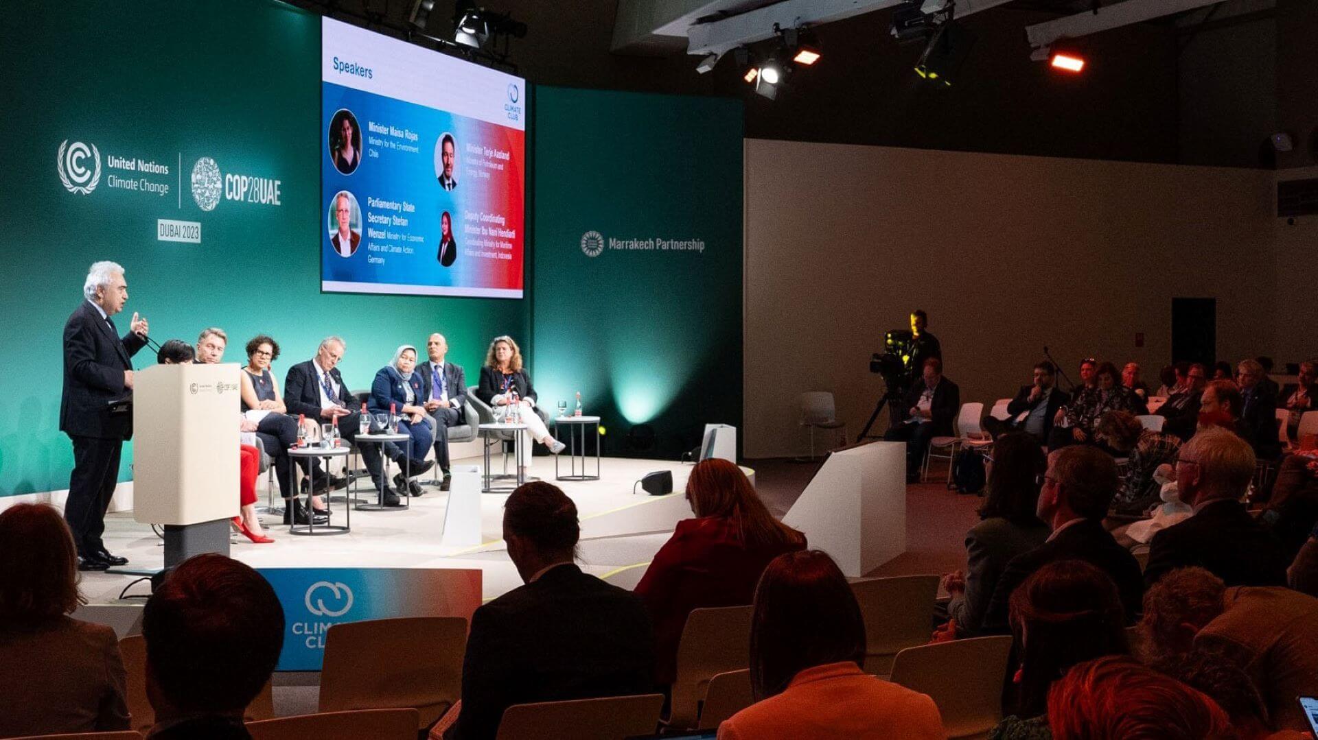 Shot from back of room, showing backs of delegates looking at stage with panel of people seated in chairs, with screen behind, and man sanding at lecturn, 