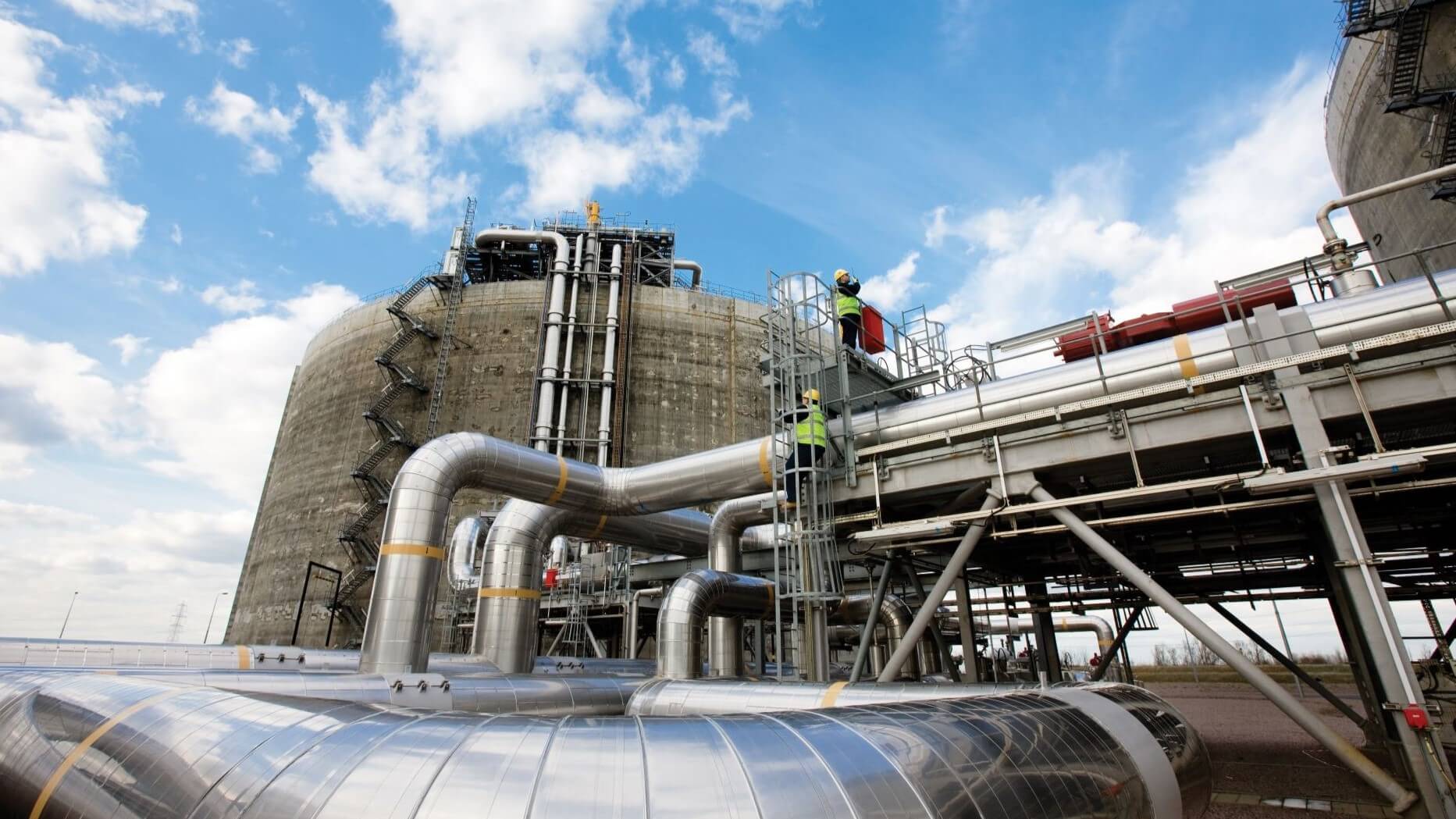 Lots of silver coloured industrial pipework running horizontally across picture, with tank in background