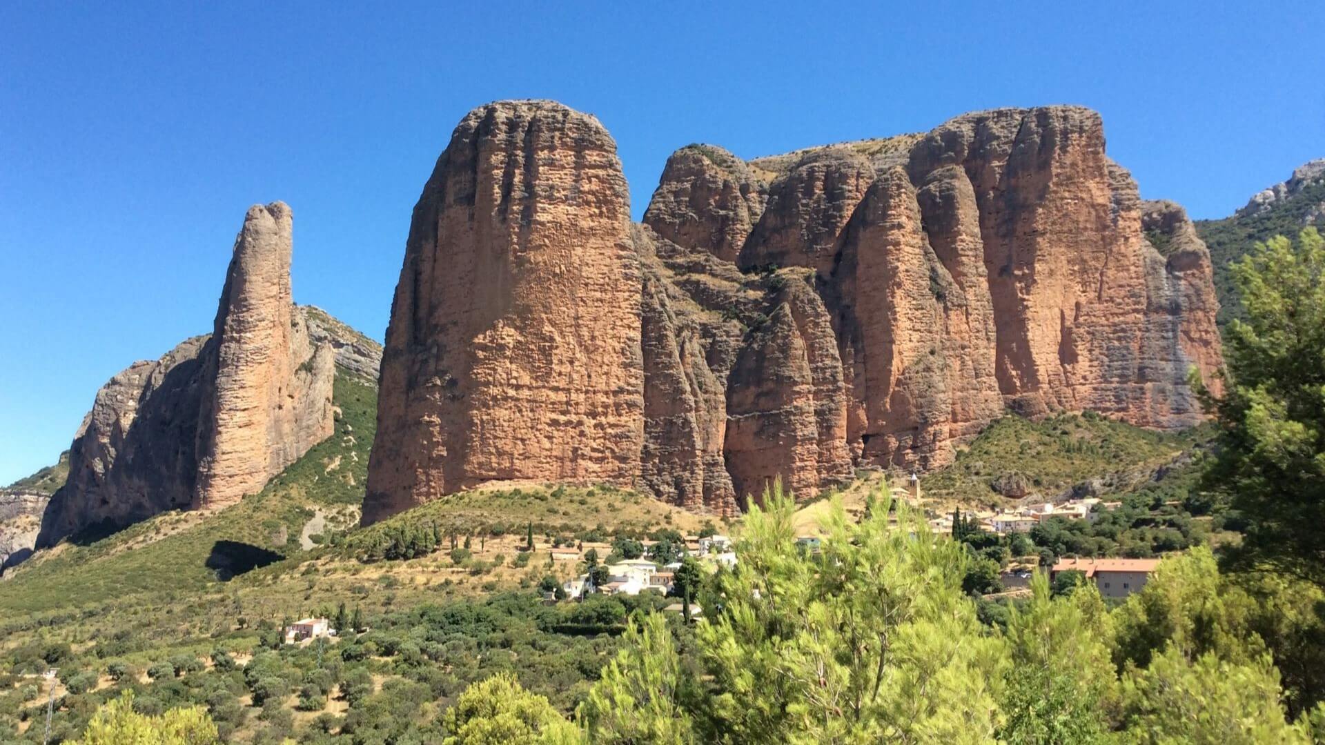 Massive striking red rock outcrop set against blue sky with green trees and undergrowth in front of rocks