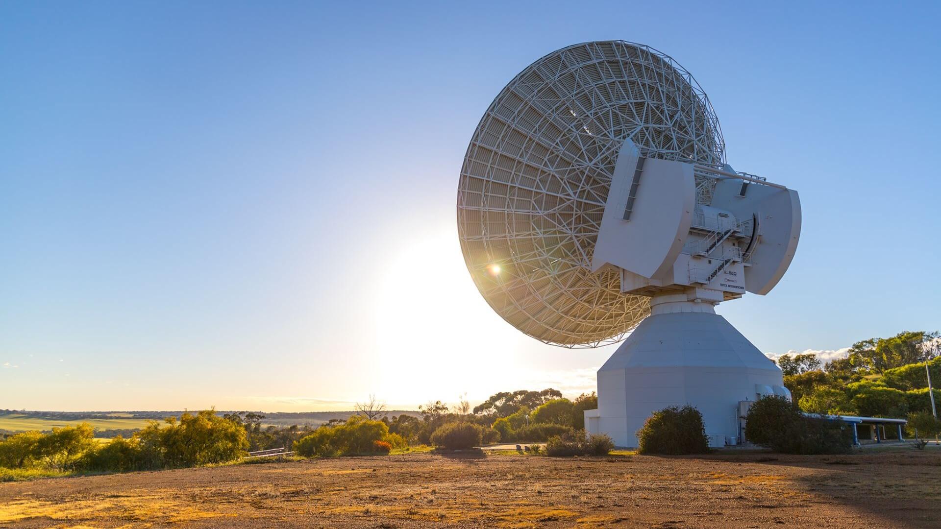 Large satellite communications disc pointing towards blue sky, with sun behind; standing in orange soil with scrubby vegetation