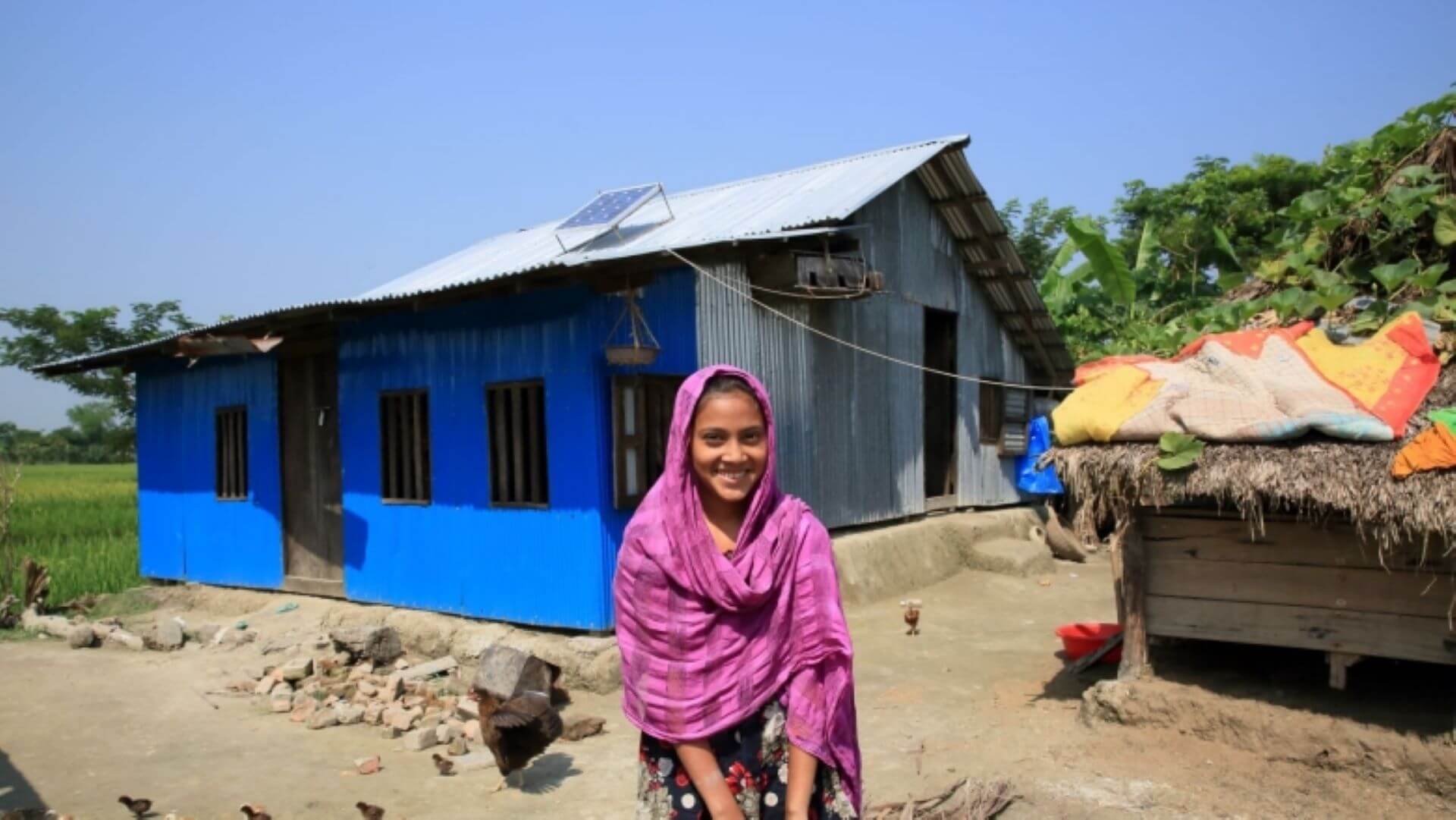 Woman in headscarf standing in front of small shack with solar panel on roof 