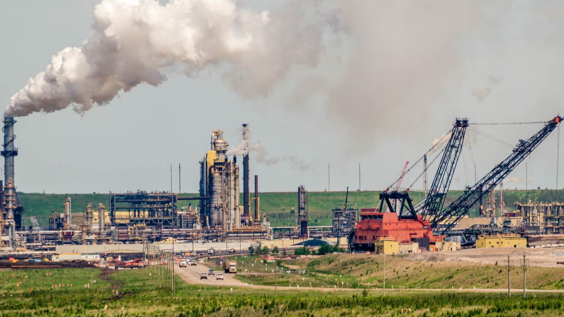 Distant view of oil sand operations showing plant and cranes and vapour cloud from stack