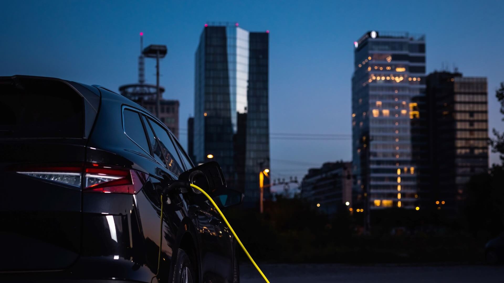 Close up of rear of electric vehicle with cable running to charger, set against night-time skyscraper skyline