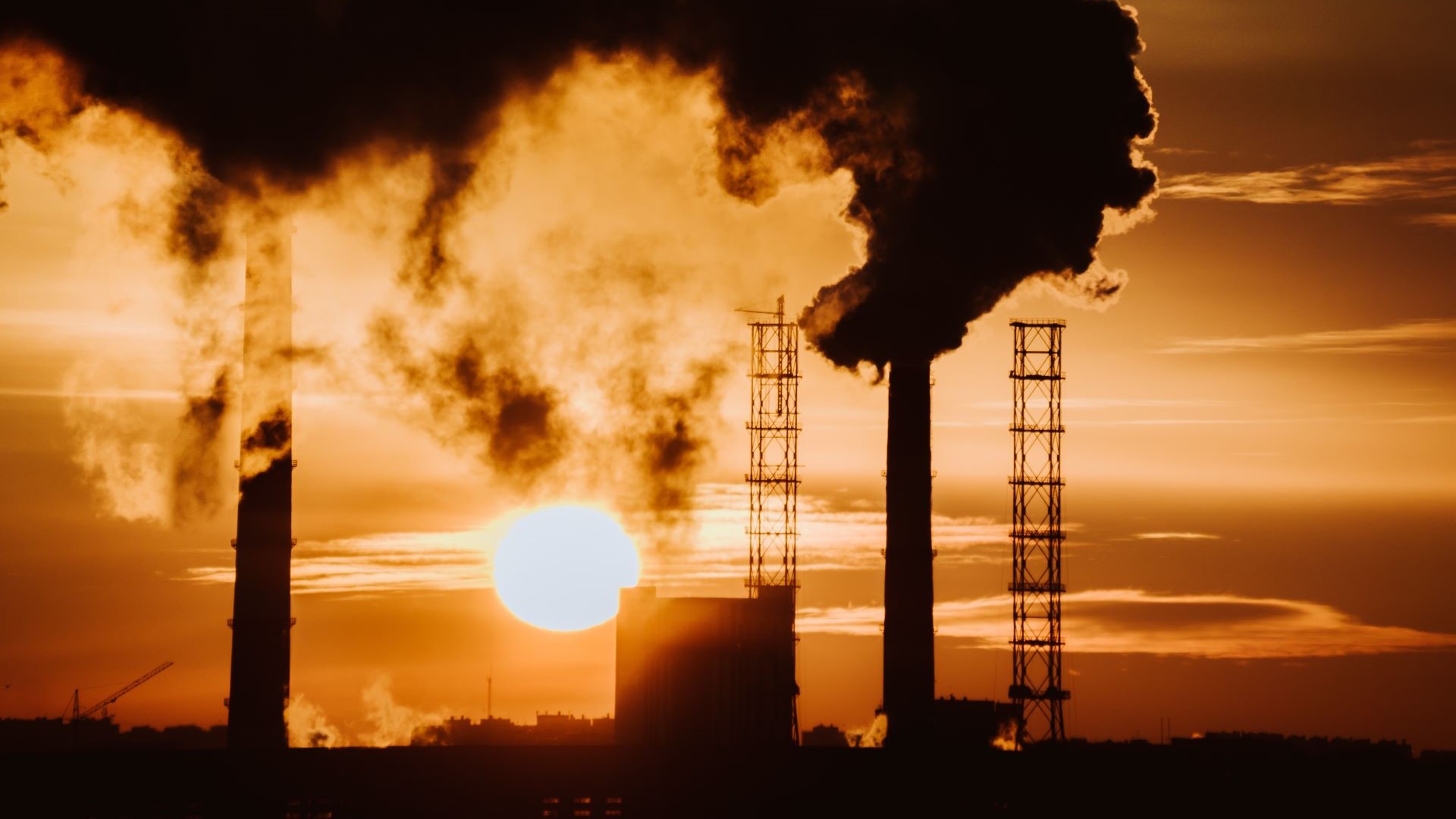 Silhouetted industrial chimneys emitting plumes of vapour set against an orange sunset sky