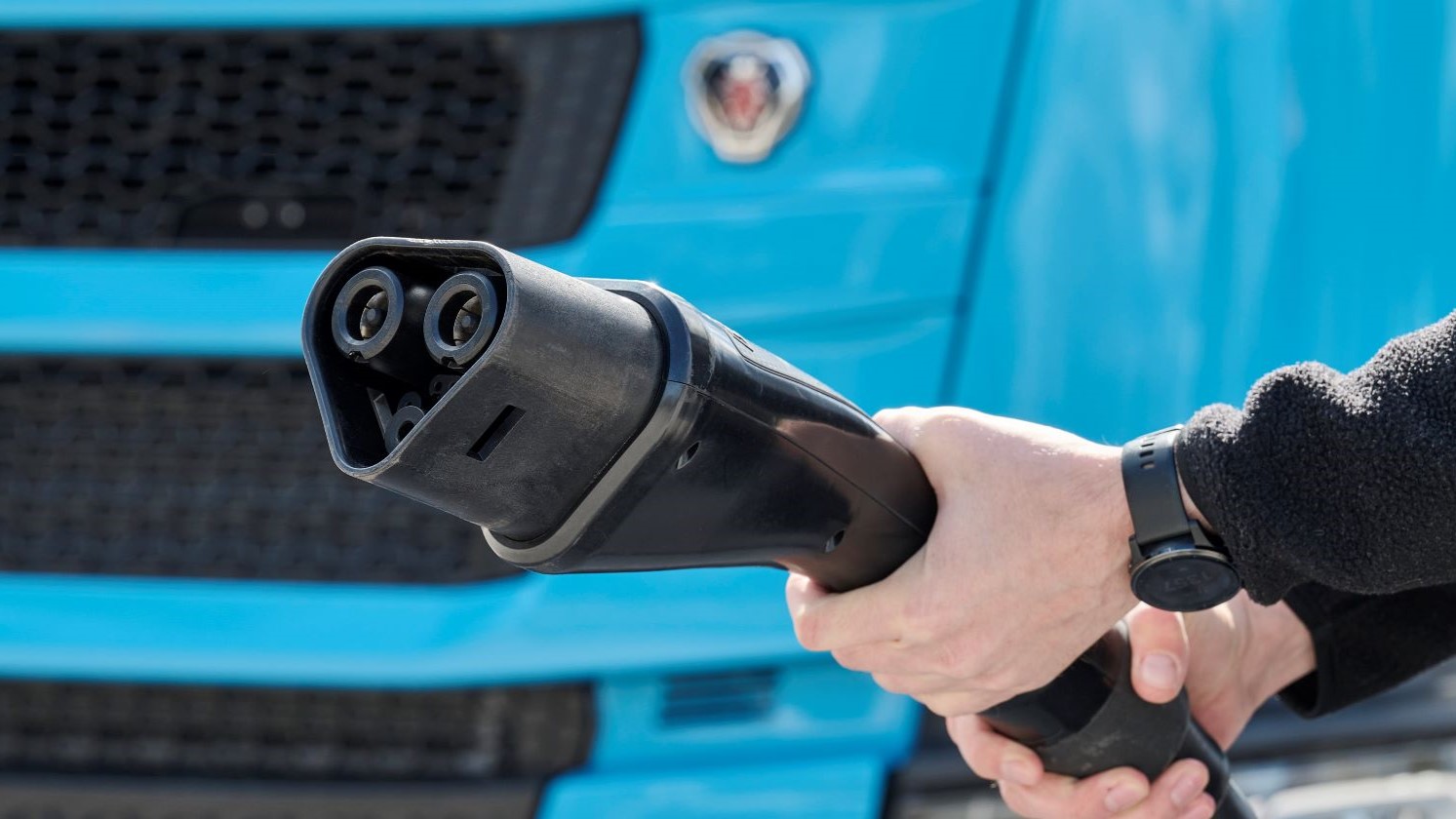 Close up of two hands gripping a large electric truck charging cable, with blue cab of truck in background