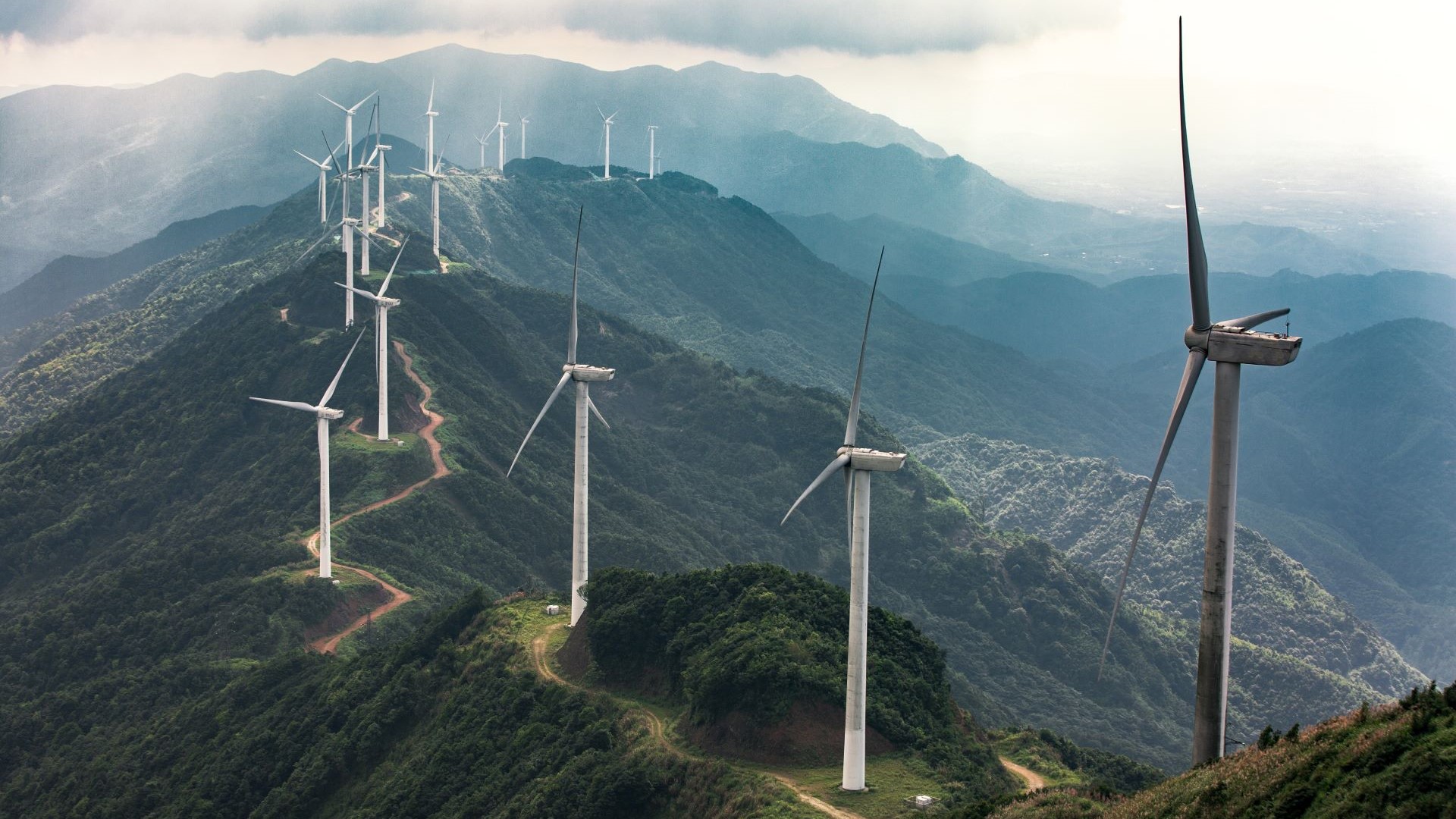 Aerial view over row of wind turbines on mountain ridge, extending into the distance