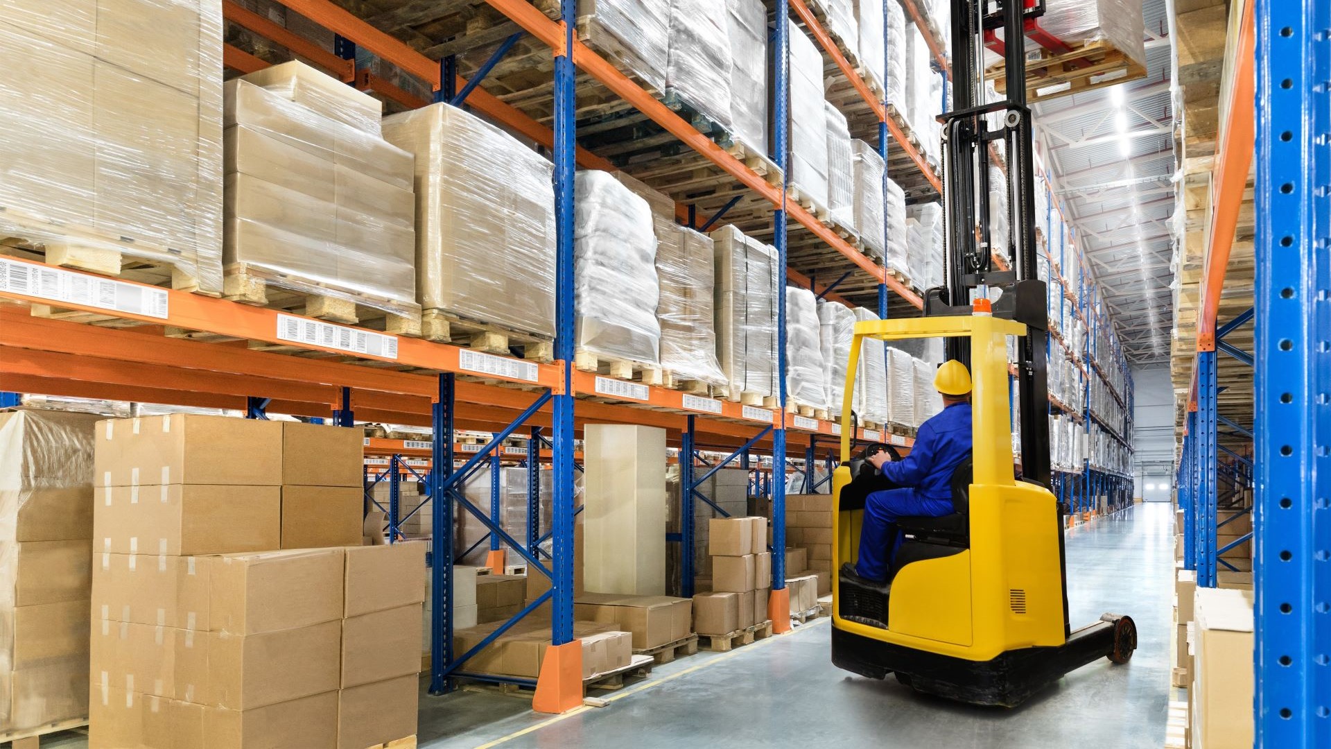 Forklift truck with forks high in air carrying a pallet along a row in a warehouse full of shelving and stacked pallets